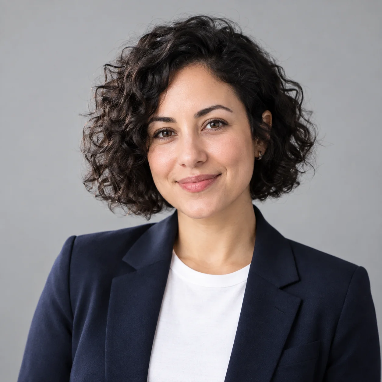 A ChatGPT headshot of a smiling woman with a chin-length curly black bob in a navy blazer and white t-shirt against a light-grey studio background